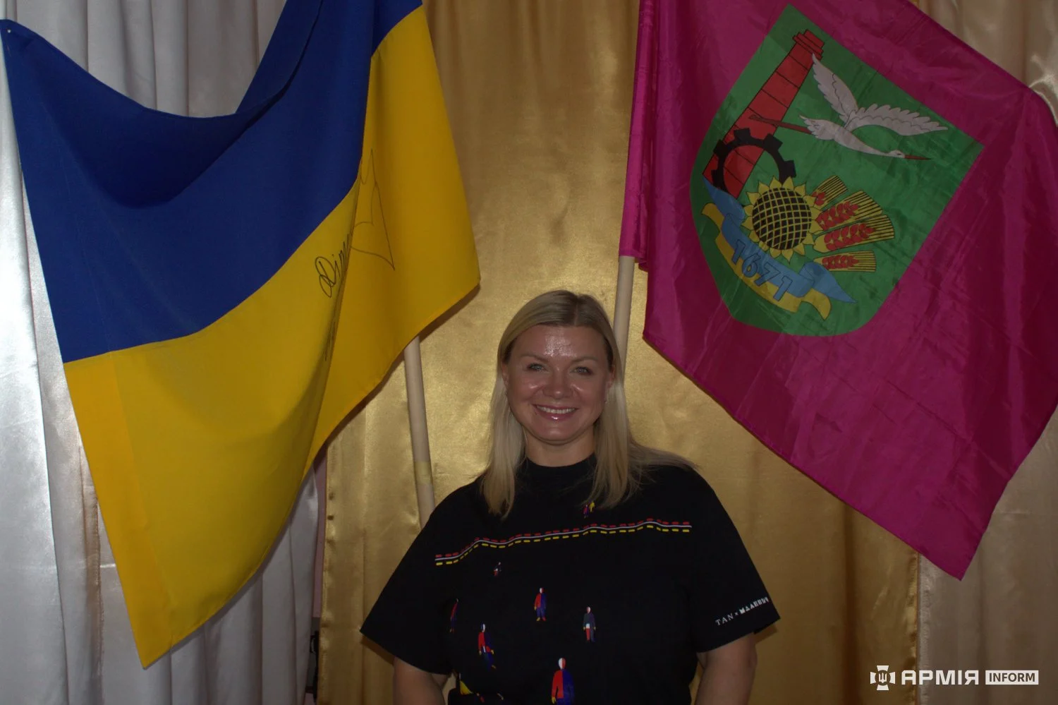 Жінка стоїть між державним прапором України та малиновим прапором із місцевою символікою. Вона дивиться перед собою й усміхається.