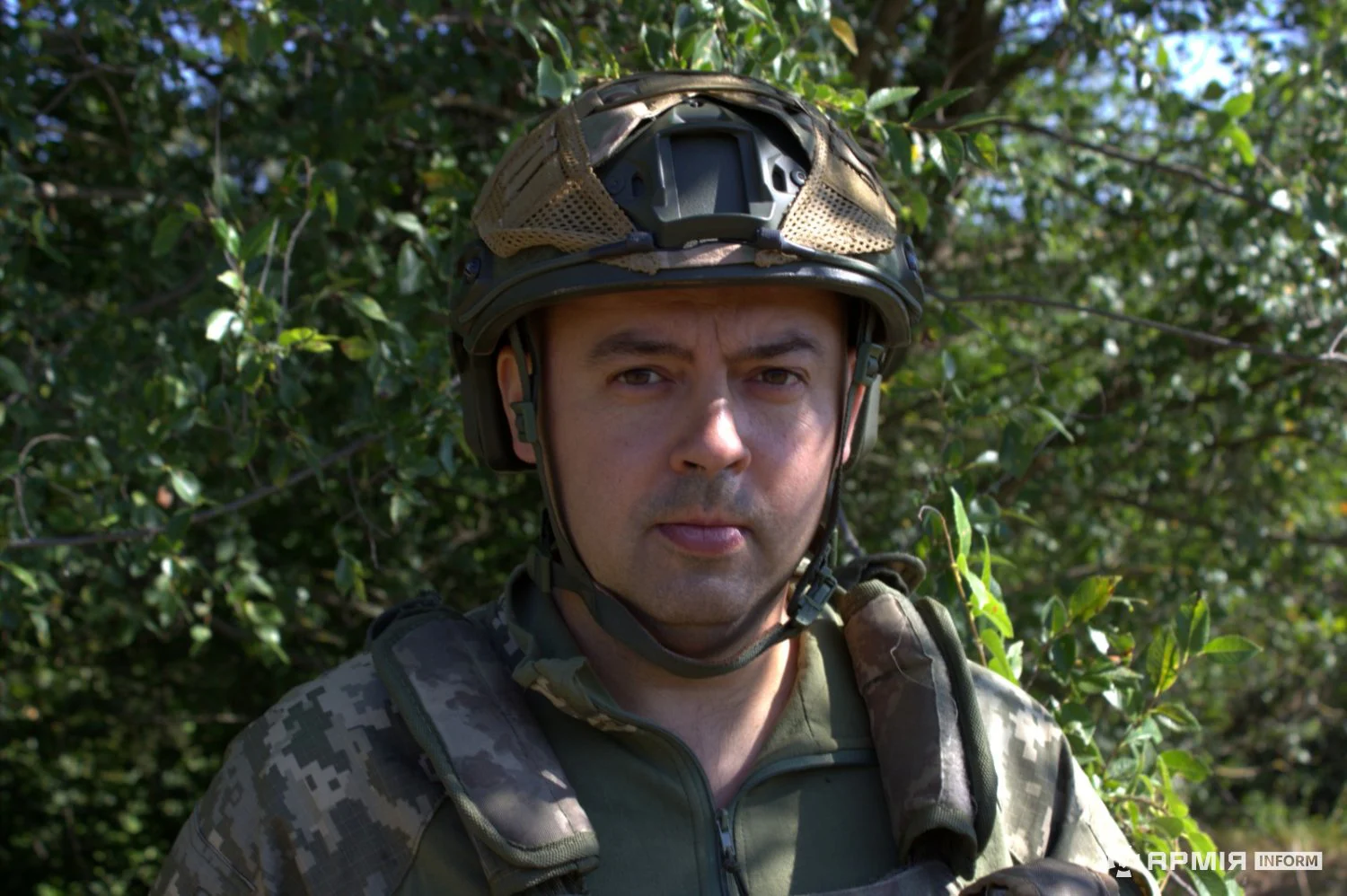 Військовий у камуфляжній формі, бронежилеті та шоломі дивиться прямо перед собою. Позаду нього ростуть дерева й густі кущі.