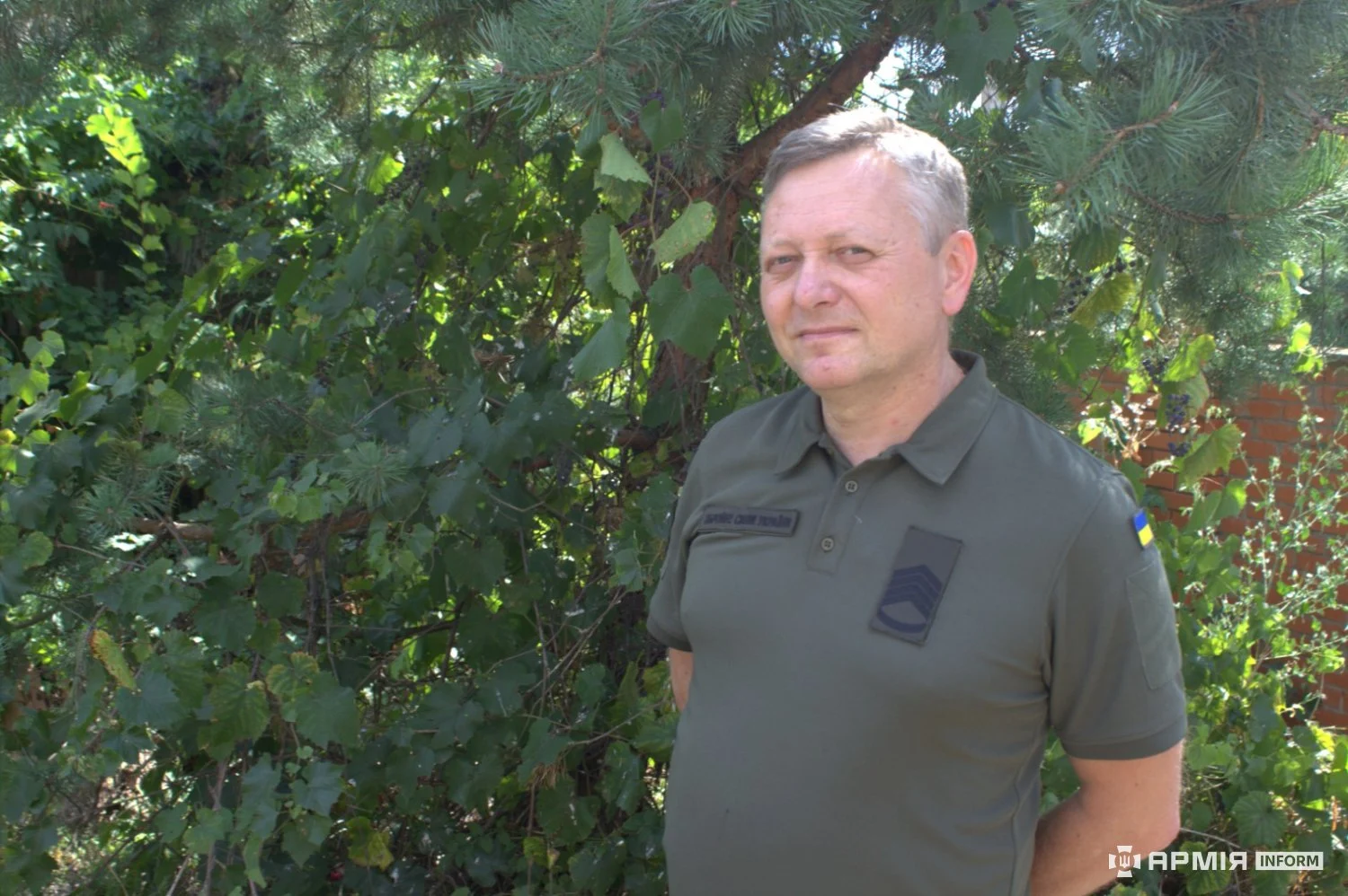 Військовий в оливковій футболці стоїть із руками за спиною біля густої зелені. На його рукаві закріплена нашивка з прапором України.