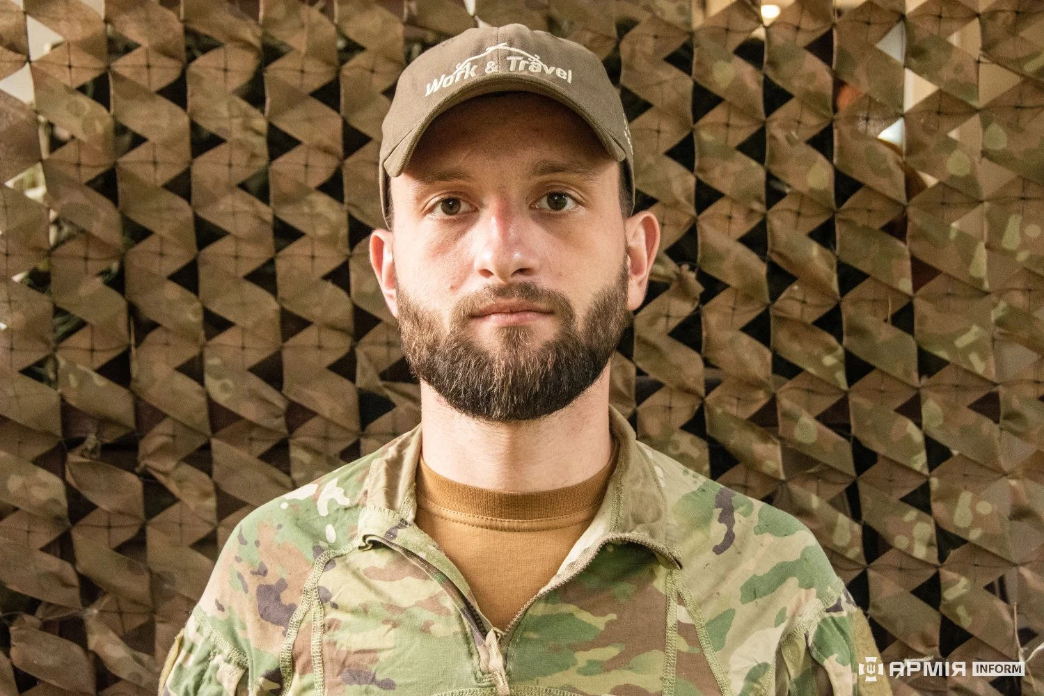 Чоловік у камуфляжній куртці та кепці дивиться в об’єктив. За його спиною натягнута маскувальна сітка.