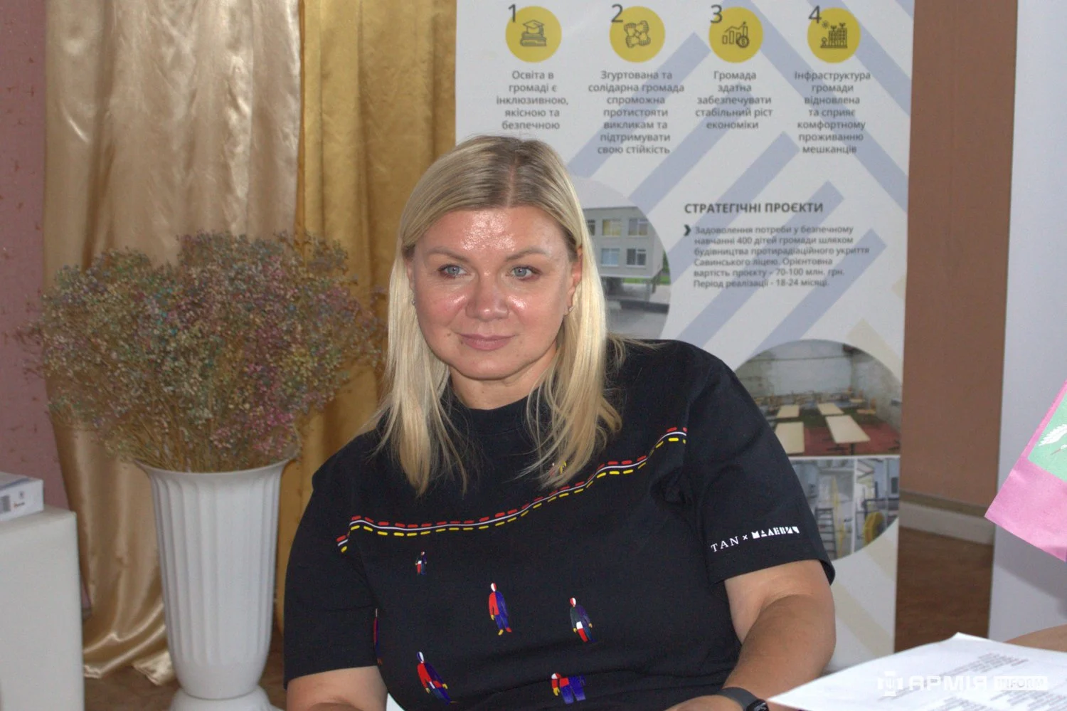 Жінка сидить за столом і дивиться перед собою. Позаду розміщений інформаційний стенд зі стратегічними пріоритетами та проєктами громади.
