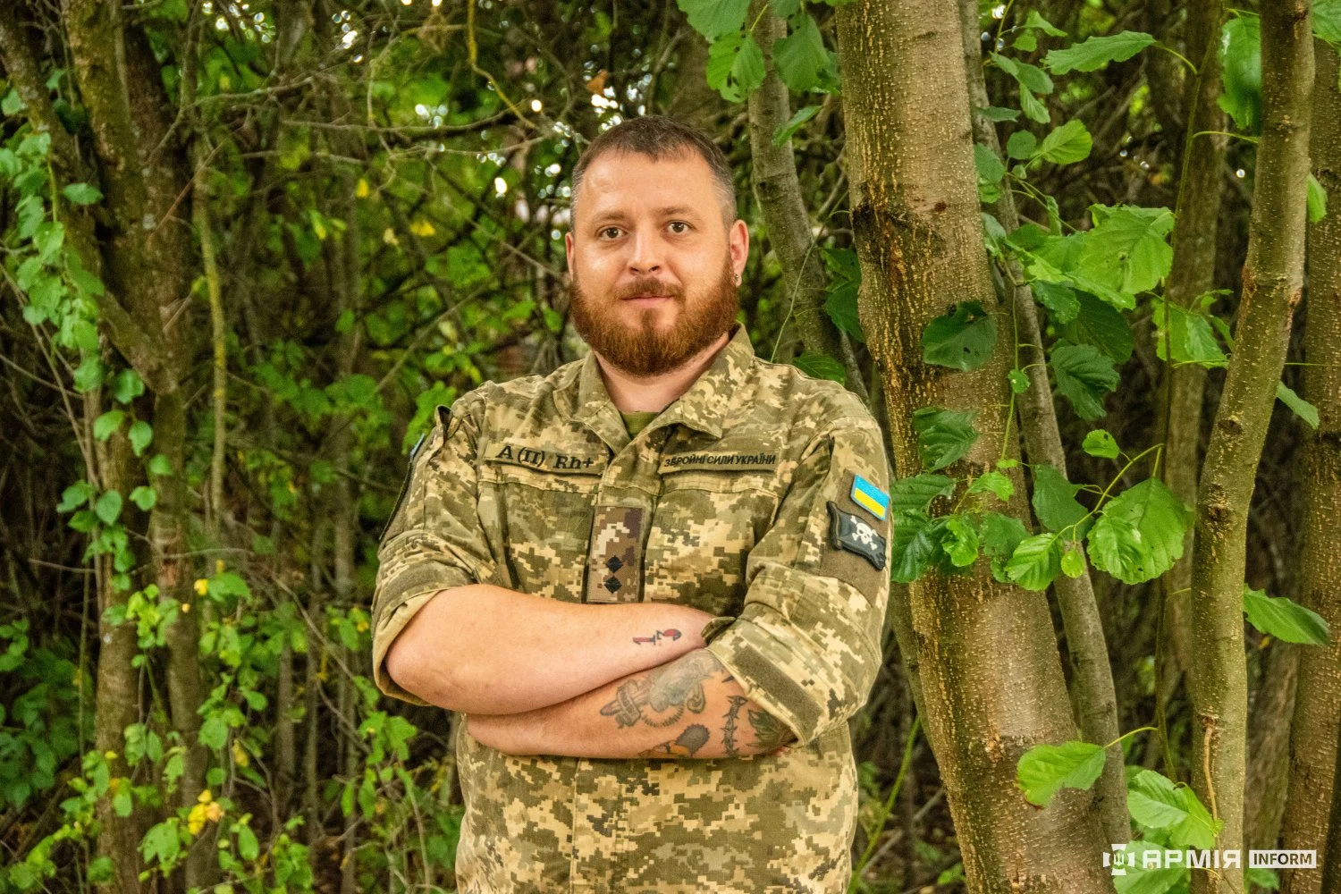 Бородатий військовий у камуфляжній формі стоїть зі схрещеними руками біля дерев. На його рукаві закріплені нашивка з прапором України та емблема, а на передпліччях є татуювання.