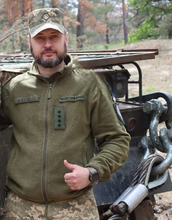Арсенал пам'яті: як зберігають спадщину артилериста Дмитра Кузнецова 9 Військовий у камуфляжній формі та зеленій куртці стоїть біля військової машини. Він дивиться в об’єктив і показує піднятий великий палець. Поруч із ним закріплені товстий трос і металеві гаки.