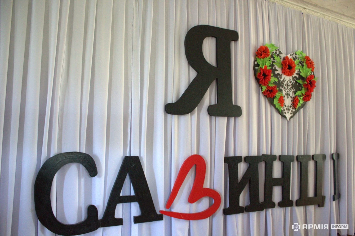 Декоративна композиція містить напис «Я Савинці» з червоним серцем замість частини слова. Поруч закріплене серце, оздоблене червоними квітами та орнаментом.