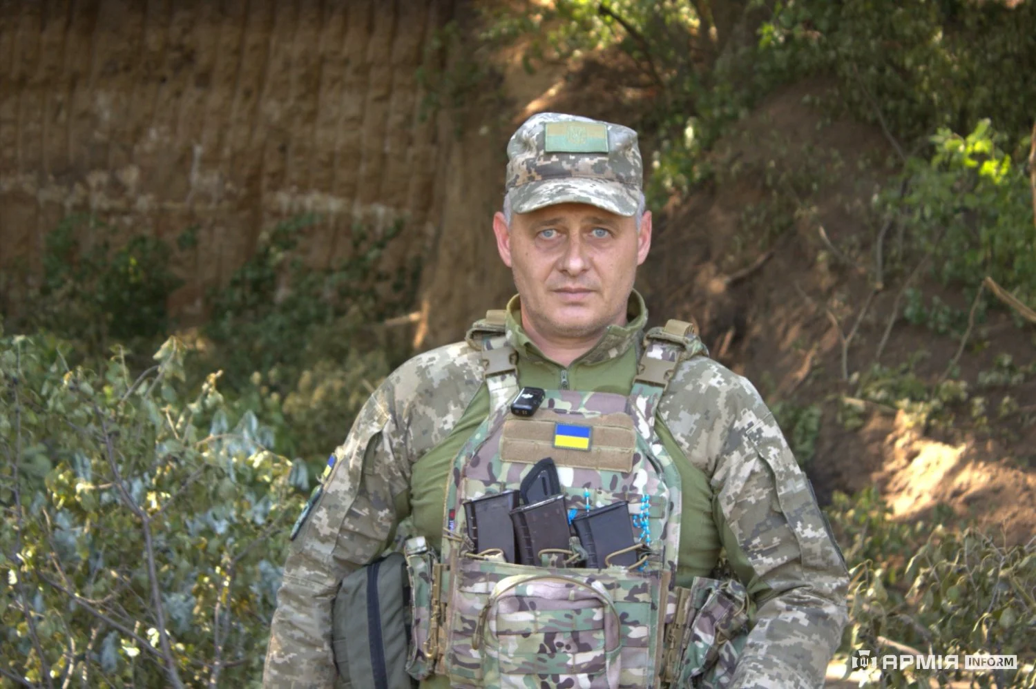 Військовий у камуфляжній формі, кепці та бронежилеті стоїть у земляному укритті. На спорядженні закріплені магазини, підсумки та нашивка з прапором України.