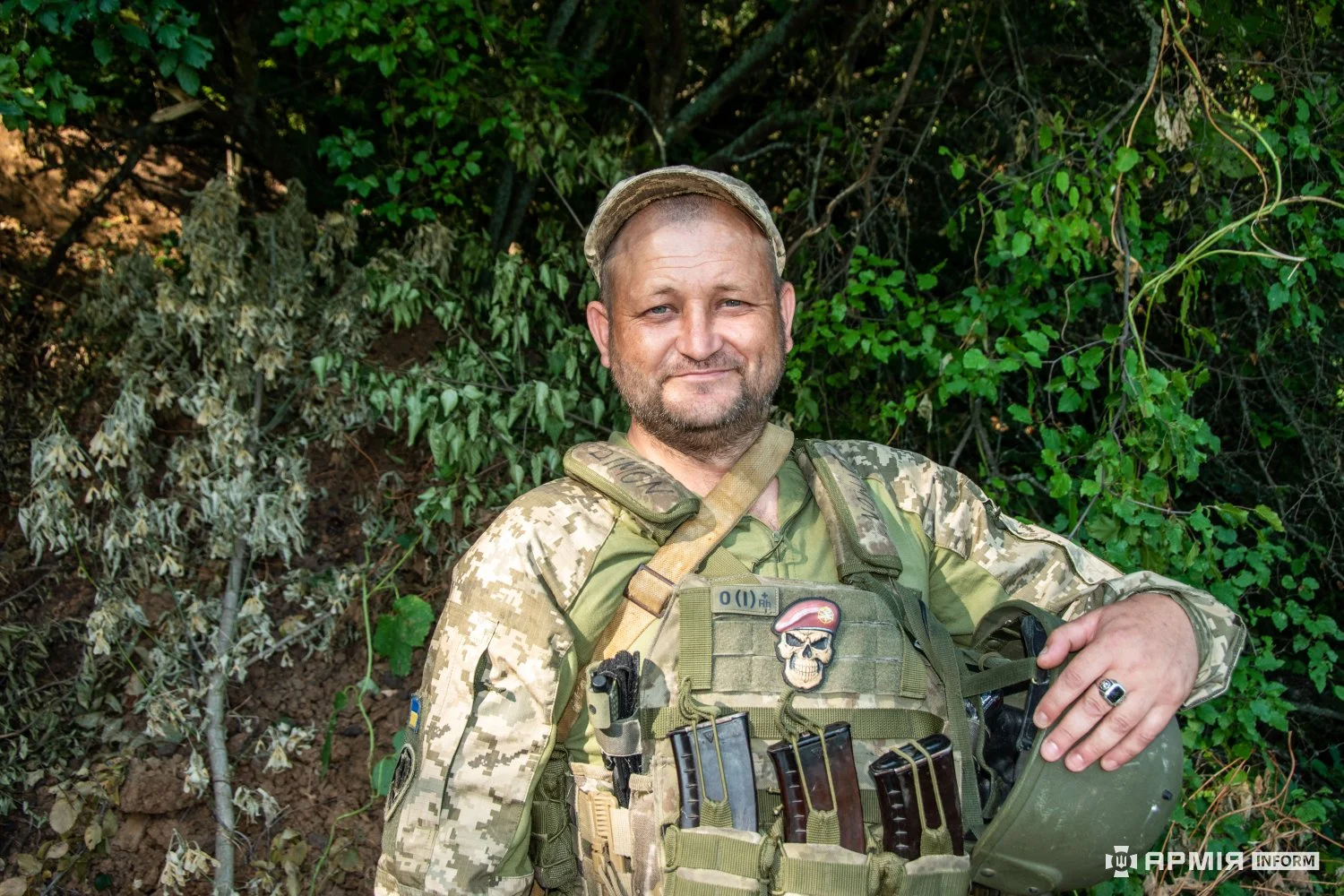 Військовий у камуфляжній формі та бронежилеті сидить біля заростей і тримає руку на шоломі. На його спорядженні закріплені магазини та нашивка із черепом у береті.