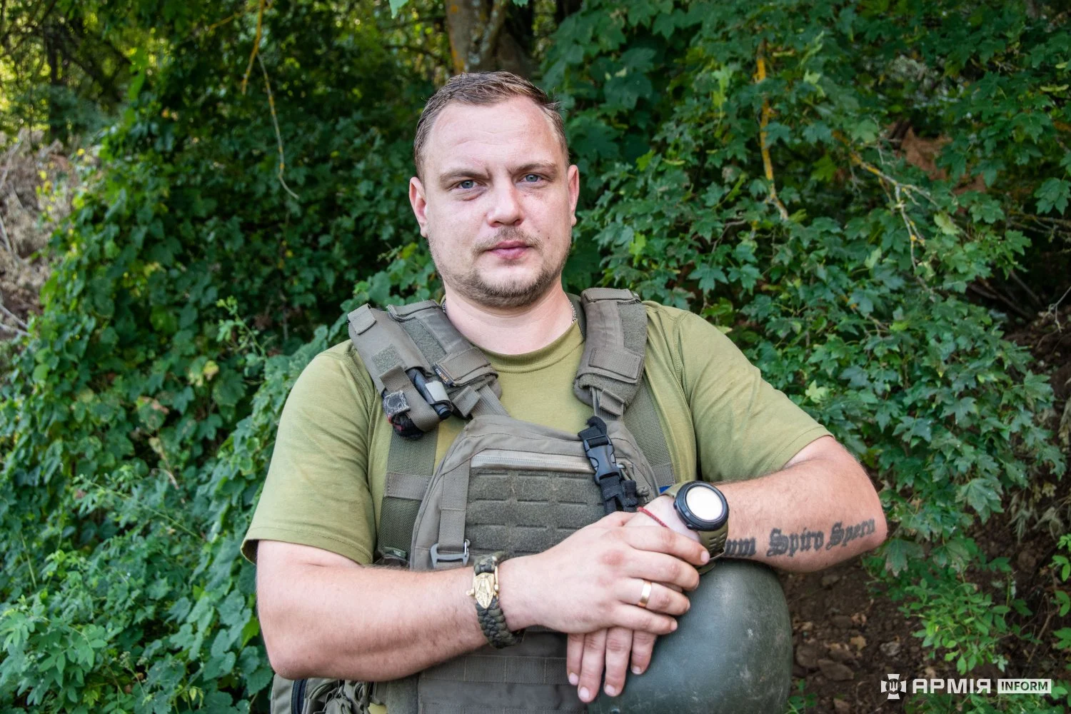 Військовий в оливковій футболці та бронежилеті спирається руками на шолом. На його передпліччі є татуювання, а позаду росте густа зелень.