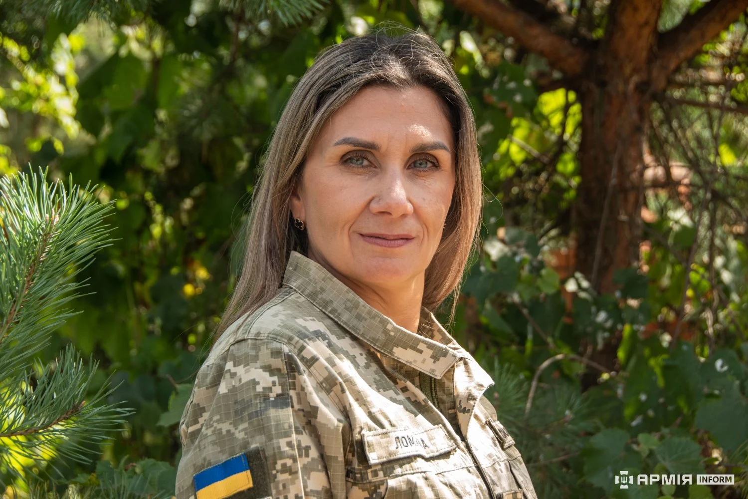 Жінка у камуфляжній формі стоїть серед хвойних дерев і дивиться в об’єктив. На її рукаві закріплена нашивка з прапором України.