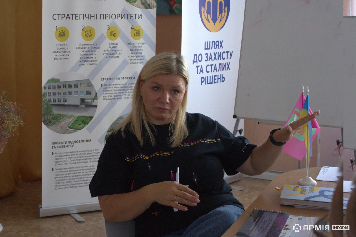Жінка сидить за столом і жестикулює правою рукою. Позаду неї розміщені інформаційні стенди зі стратегічними пріоритетами та два настільні прапори.
