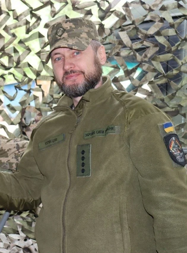 Арсенал пам'яті: як зберігають спадщину артилериста Дмитра Кузнецова 10 Військовий у зеленій куртці та камуфляжному кашкеті стоїть біля маскувальної сітки. Він дивиться в об’єктив, а на його рукаві закріплені нашивки, зокрема синьо-жовта.