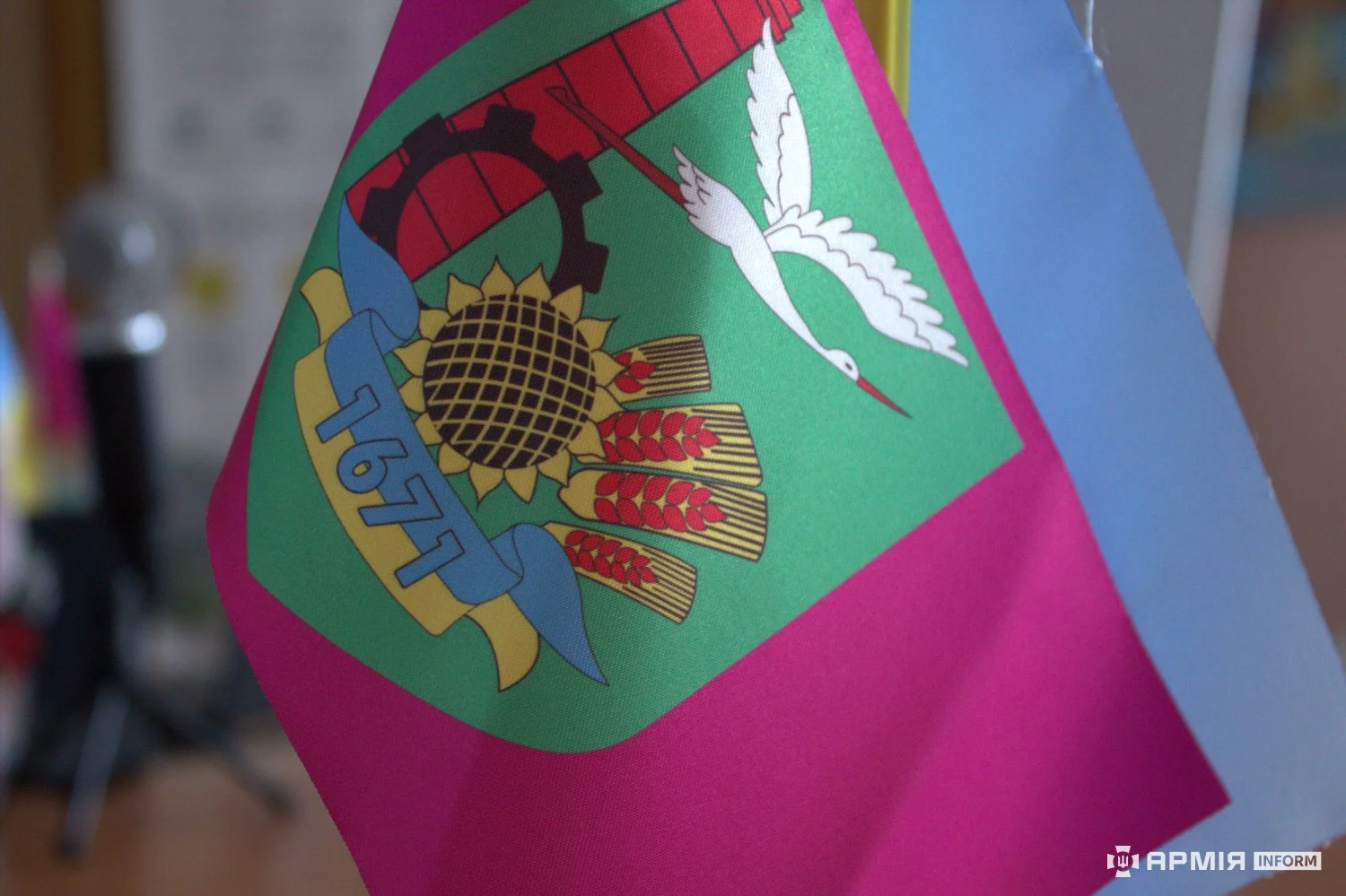 Малиновий прапор містить емблему із соняшником, колосками, птахом, промисловим символом і числом «1671». Позаду частково розташований блакитний прапор.