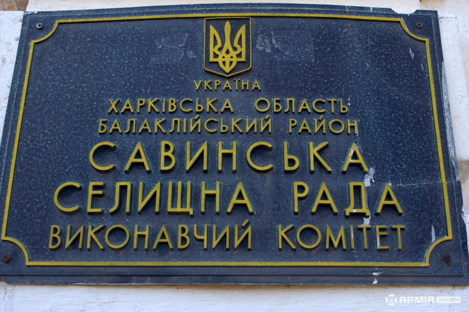 Табличка містить державний герб України та напис «Харківська область, Балаклійський район, Савинська селищна рада, виконавчий комітет». Літери й декоративна рамка виконані золотистим кольором на темному тлі.